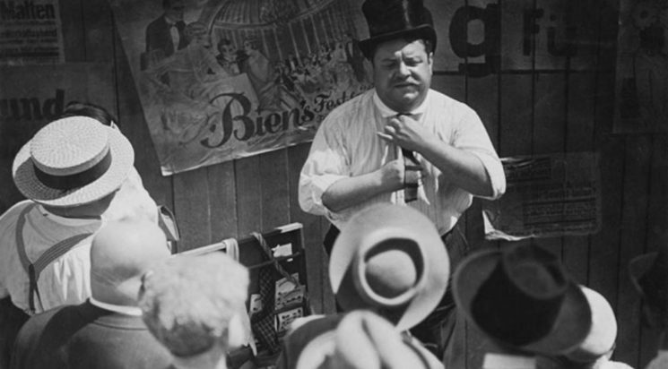 Image en noir et blanc tirée du film Berlin Alexanderplatz de Phil Jutzi : un homme en chemise et haut chapeau présentant des cravates à un petit groupe vu de dos