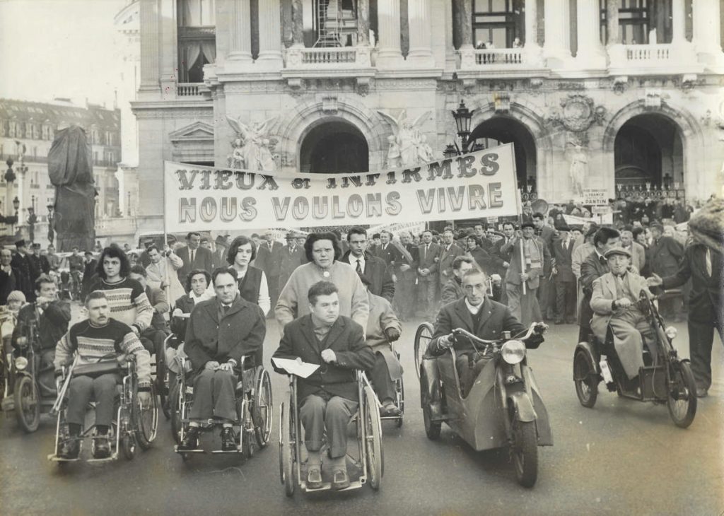Photographie en noir et blanc : au premier plan, une dizaine de personnes en fauteuil roulant ou tricycle, manifestant devant une banderole « Vieux et infirmes, nous voulons vivre », avec un large groupe de manifestants debout à l'arrière-plan.