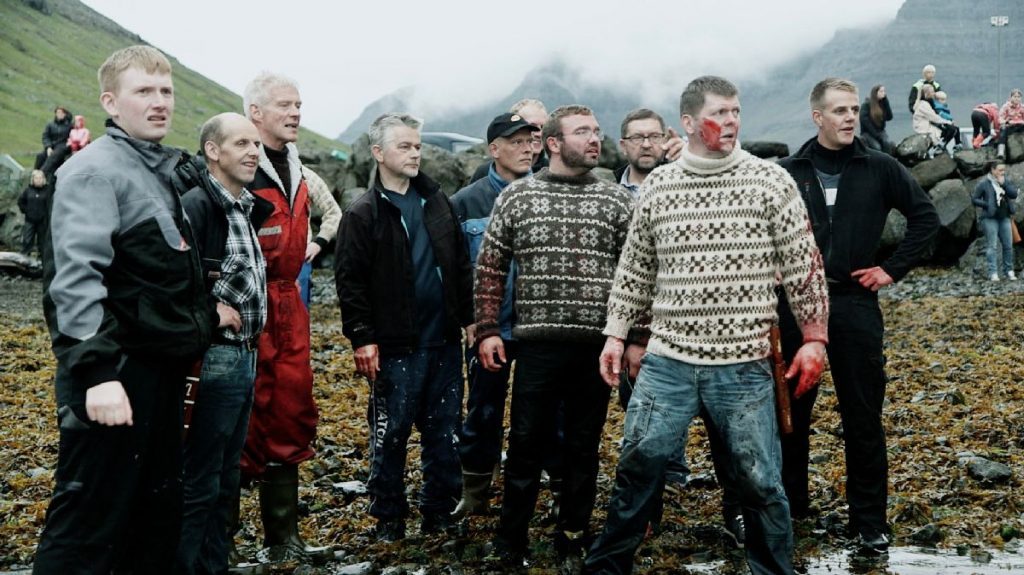 Dix hommes debout sur une plage d'algues et de cailloux, en tenue de pêche ou d'hiver. Ils regardent tous dans la même direction, à droite de la caméra. Trois d'entre eux ont du sang sur les mains, un quatrième également sur les joues.