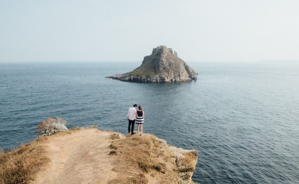 Un couple debout, de dos, sur falaise en bord de mer avec, à l'arrière-plan, un îlot rocheux.