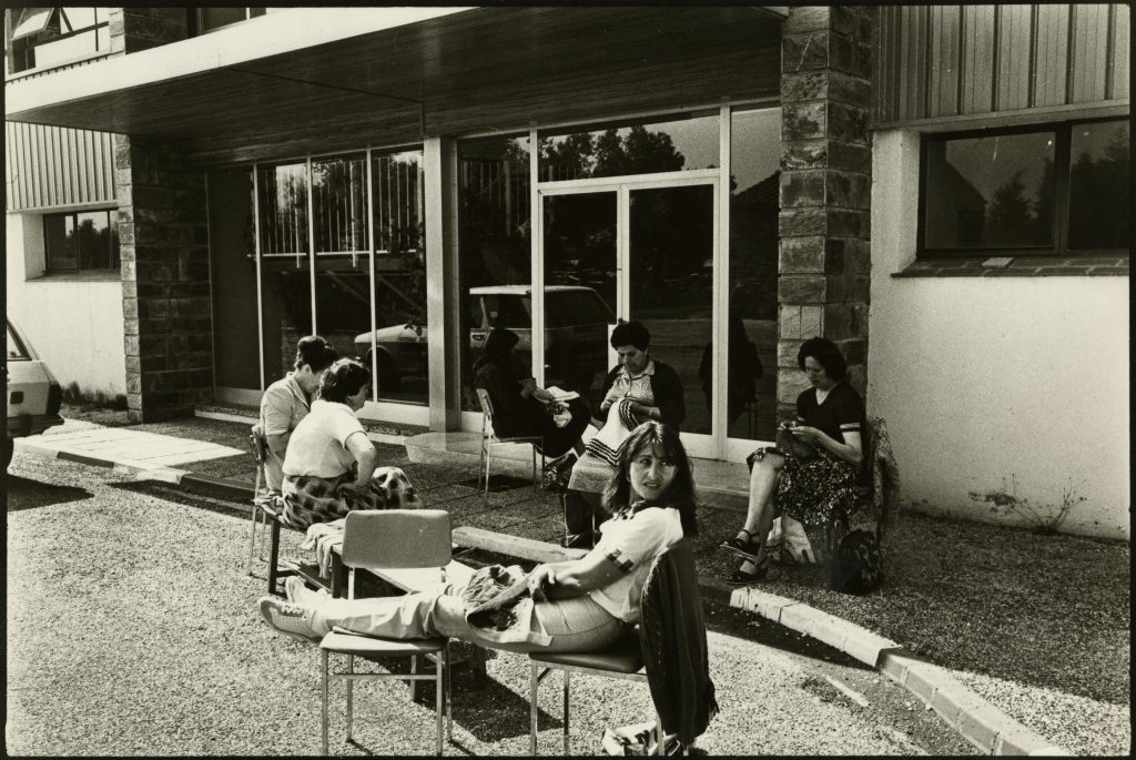 Occupation d'usine en 1981. Les femmes sont assises devant l'entrée, occupées par des travaux d'aiguille.