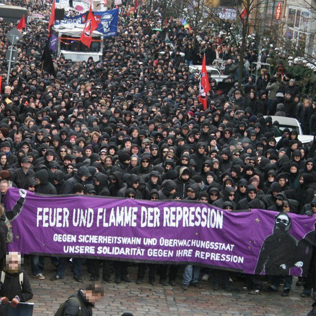 Manifestation de personnes habillées en noir avec bandeauet capuches sur le visage.