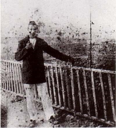 Photo d'archive en noir et blanc très dégradée. Autoportrait d'Arthur Rimbaud, de face, il porte un pantalon blanc et une veste de marin, et est appuyé sur une barrière.