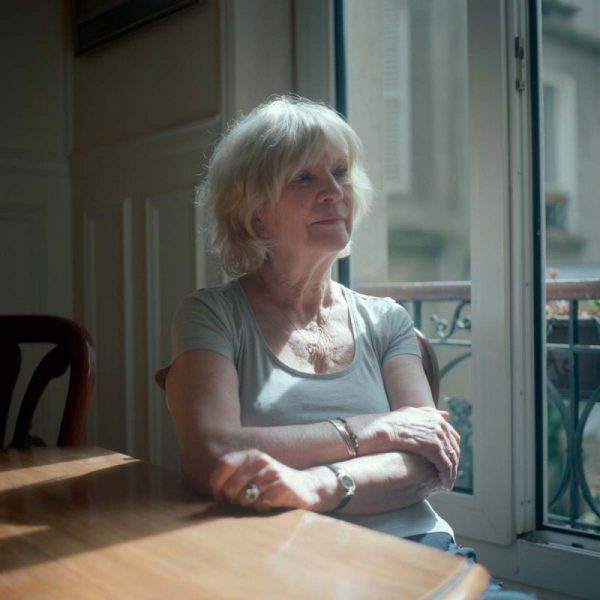 Photo couleur. Femme d'une soixantaine d'années, assise près d'une fenêtre qui laisse passer un rayon de soleil. De trois quarts, sa main repose sur une table