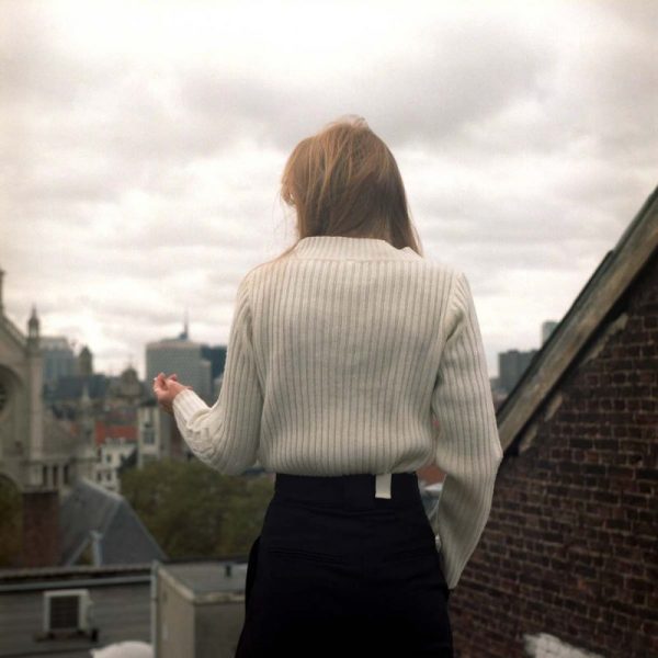 Photo couleur d'une femme de dos, fumant une cigarette sur une terrasse. Au second plan le ciel couvert et les toits de la ville