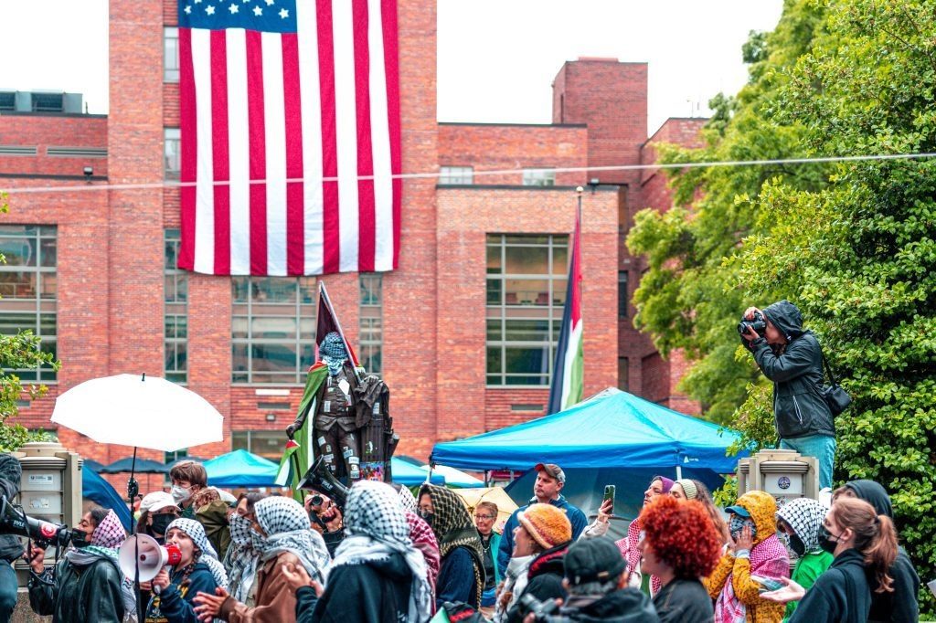 Photo montrant une manifestation de protestation anti-israélienne sur le campus de l’Université Georges Washington, à Washington DC, en 2024. Certains manifestants portent un keffieh. Au second plan, un bâtiment de l'université arborant le drapeau des États-Unis.