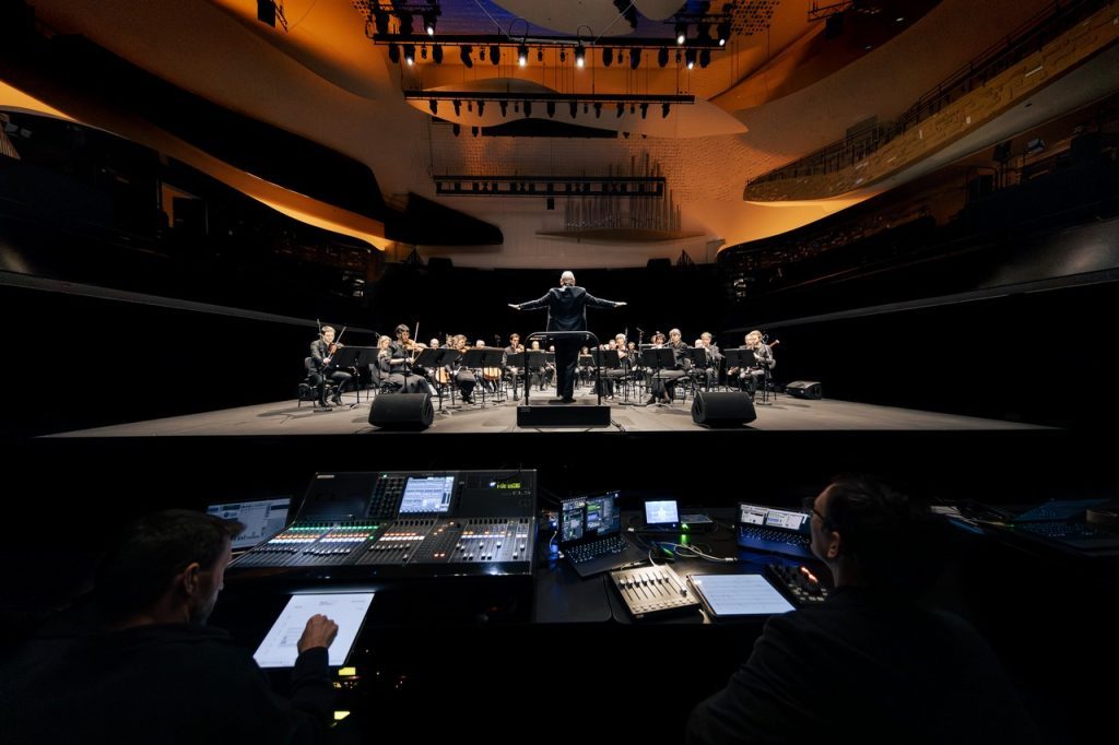 Photo couleur, prise à la Philharmonie de Paris le 6 janvier 2025. Au premier plan les ingénieurs du son et leur machines; Au deuxième plan, en hauteur l'Ensemble intercontemporain et le chef d'orchestre Pierre Bleuse.