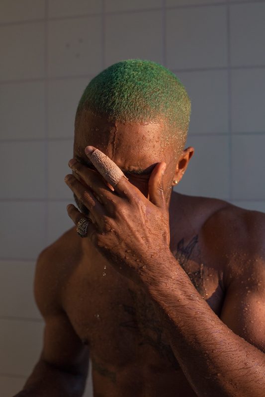 Frank, in the shower, de Wolfgang Tillmans, réalisé en 2015, est un portrait puissant capturant un homme torse nu sous une douche carrelée de blanc. L’homme, aux cheveux teints en vert vif, est saisi dans un moment d'intimité : il se couvre partiellement le visage de la main, les gouttes d’eau perlant sur sa peau sombre et musclée. Un bandage clair entoure l’un de ses doigts, ajoutant une note de fragilité à la force de sa posture. La lumière naturelle caresse son corps, soulignant la texture humide de sa peau et révélant subtilement un tatouage sur son torse. L’expression du sujet, bien que partiellement dissimulée, suggère une émotion profonde, entre pudeur, recueillement et intensité intérieure. Par cet instantané, Tillmans explore la vulnérabilité masculine et la beauté brute, dans un dialogue nuancé entre présence physique et intériorité émotionnelle.