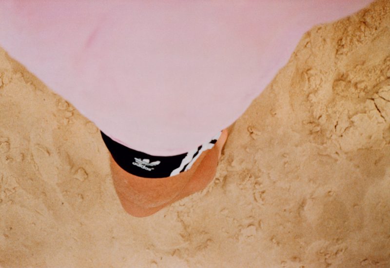 Lacanau (self) de Wolfgang Tillmans, réalisée en 1986, propose une vue subjective et intimiste ; un moment de détente à la plage. Photographiée depuis un angle plongeant vers le sol, l’image montre les jambes nues de l’artiste partiellement couvertes par un short noir Adidas, surmonté d'un t-shirt rose pastel qui envahit la partie supérieure du cadre. Le sable clair, parsemé d’empreintes et de textures naturelles, occupe tout l’arrière-plan. L'absence de repères directs (horizon, mer) recentre l’attention sur l’immédiateté de l'expérience corporelle et sensorielle. L'image évoque une sensation d’ancrage et de simplicité, traduisant l’esprit de liberté, de chaleur estivale et d’introspection douce qui caractérise souvent le travail de Tillmans. Cette photographie témoigne aussi d’une approche documentaire et autobiographique précoce, où l’instant personnel devient universel.