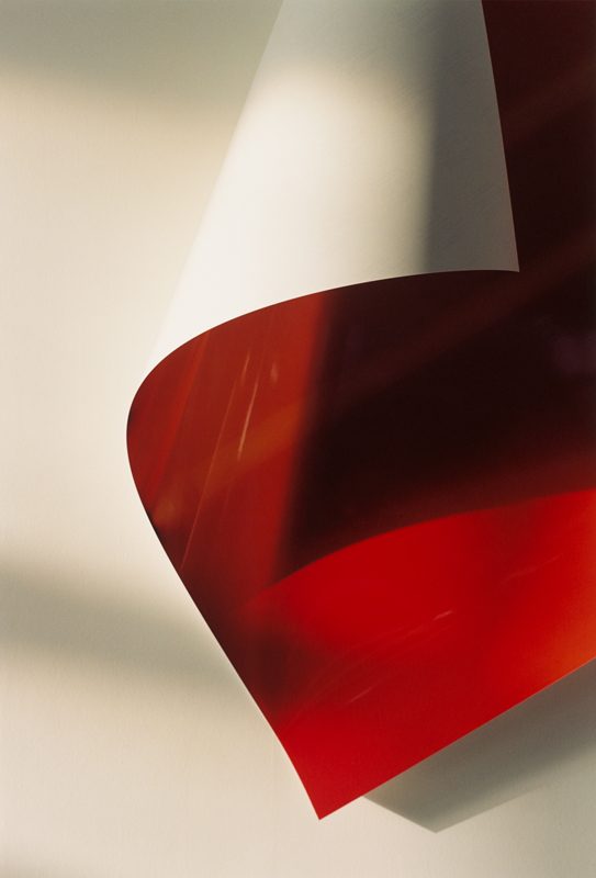 Paper drop 22, de Wolfgang Tillmans, réalisée en 2001, présente une composition épurée où une feuille de papier se déploie en une élégante courbe suspendue. La partie supérieure de la feuille reste blanche, tandis que sa face inférieure, d’un rouge éclatant et légèrement translucide, capte et reflète la lumière ambiante. Les jeux d’ombres portées sur le mur neutre renforcent la tridimensionnalité du papier, accentuant l’illusion d’un mouvement délicat figé dans l’instant. Les lignes douces et les transitions subtiles entre les tons créent une esthétique à la fois minimaliste et sensuelle. Tillmans explore ici les propriétés physiques du papier — sa légèreté, sa flexibilité, sa capacité à modeler l'espace — tout en transcendant la nature ordinaire de l’objet pour en révéler une beauté sculpturale. La maîtrise de la lumière naturelle joue un rôle clé, conférant à la scène une douceur presque tactile.