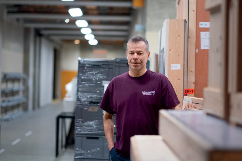 Wolfgang Tillmans est photographié dans un espace logistique du Centre Pompidou, au milieu des caisses de transport destinées aux œuvres d'art. Vêtu d’un t-shirt bordeaux marqué « Démé à Paris », il adopte une posture naturelle, une main dans la poche, l’autre posée légèrement sur une caisse en bois. Son regard, serein et attentif, traduit une implication directe dans les opérations de montage de son exposition. L’environnement industriel, avec ses caisses en bois étiquetées « Fragile », ses rayonnages métalliques et ses lignes de fuite marquées par les plafonniers alignés, évoque l'envers discret mais fondamental du monde muséal. Les tons neutres de béton brut, bois clair et emballages plastiques participent à instaurer une atmosphère méthodique, révélant la dimension matérielle de la création et de l'exposition artistique.