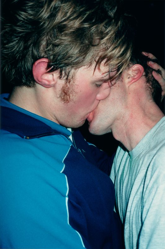 The Cock (Kiss)" de Wolfgang Tillmans, réalisée en 2002, saisit avec intensité l’instant brut d’un baiser passionné entre deux hommes. La scène se déroule dans une atmosphère nocturne, probablement dans un club ou un espace festif. Les deux figures, plongées dans l'intimité de leur étreinte, occupent presque tout le cadre, accentuant la force émotionnelle de l’image. L'un porte une veste de sport bleu vif, l’autre un t-shirt gris clair, contrastant légèrement par leurs postures et carnations. Les détails — mèches ébouriffées, rougeurs sur la peau, tension des mains agrippant la nuque — renforcent l’authenticité et l’urgence du moment vécu. La lumière directe du flash accentue la texture de la peau et donne à l'image une immédiateté quasi viscérale. À travers ce cliché, Tillmans explore la tendresse, la sexualité, la vulnérabilité et la liberté, brisant les normes traditionnelles de représentation de l’amour queer et affirmant une esthétique radicalement honnête et inclusive.