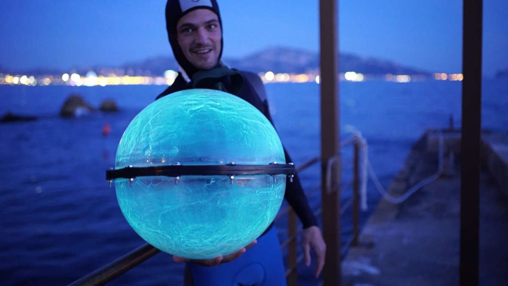 Jeremy Bruguidou en tenue de plongée, une de ses sphères bioluminescentes à la main. La sphère en gros plan fait environ 60 cm de diamètre