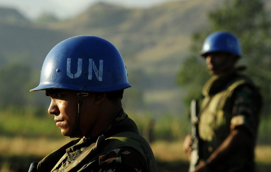 Deux membres des forces de sécurité des Nations Unies, venus du Népal, sécurisent une zone de largage avant un parachutage de palettes. Gros plan sur la tête casquée de bleu du premier soldat. En fond, flouté, le second, équipé d'une arme à feu.