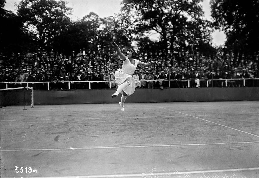Photo d'archive en noir en blanc, montrant la joueuse de tennis Suzanne Lenglen sur le court dans une posture acrobatique, dans sa robe blanche dessinée par Jean Patou.
