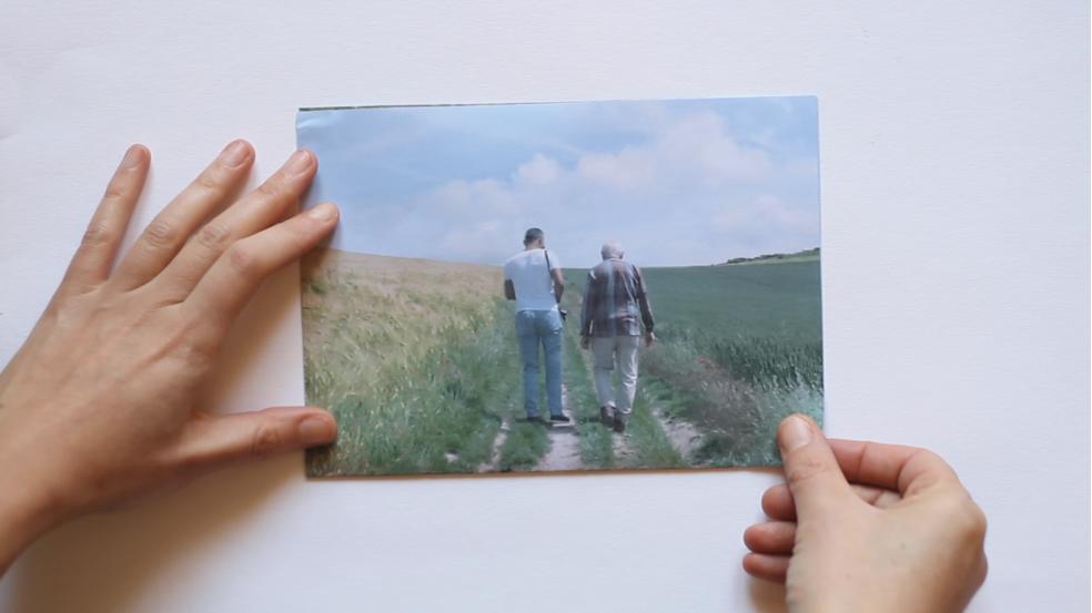 Une photographie de deux hommes marchant de dos dans un champ, tenue par deux mains contre un mur