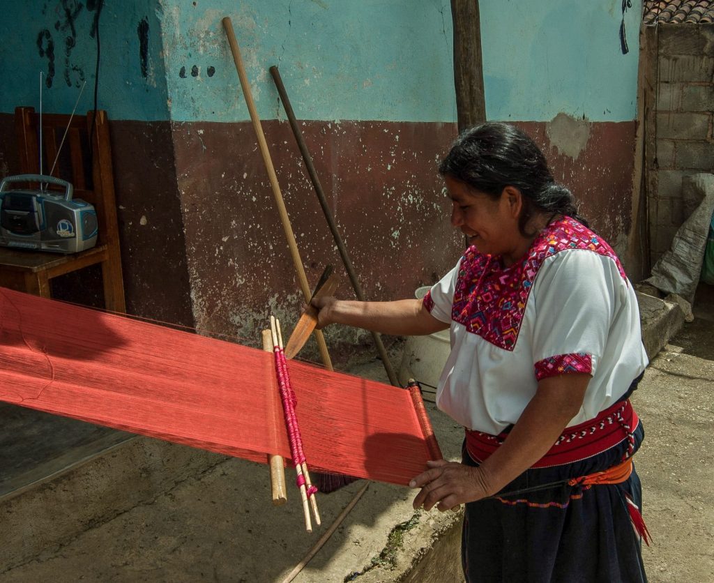 Photo couleur. Une tisseuse, portant le huipil, utilise un telar de cintura, métier à tisser à sangle dorsale, pour réaliser un tissu de huipil.