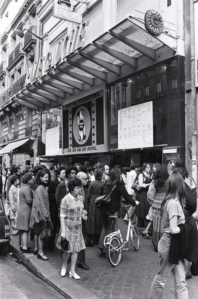 Des femmes devant le cinéma Gaumont - Rive gauche, durant le festival. L'affiche du festival international indique la date : du 23 au 29 avril. Photographie noir et blanc