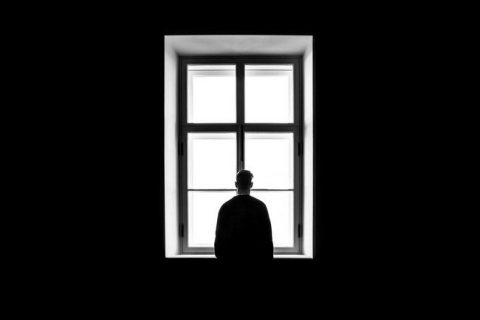 Un homme seul se tient immobile dans une pièce sombre. Debout, devant une haute fenêtre, il regarde à l'extérieur mais on ne devine rien à travers les vitres blanches.