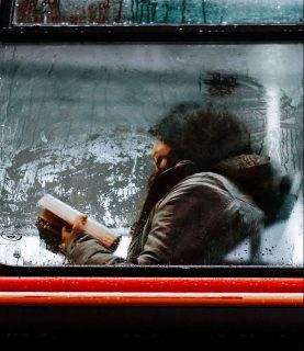Derrière une vitre embuée, sur laquelle coulent des gouttes de condensation, une femme portant manteau, écharpe et bonnet est en train de lire un livre, assise sur un siège. Elle est vraisemblablement dans un bus.