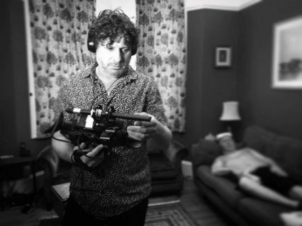 Le cinéaste Marc Isaacs, une caméra à la main dans un salon. Derrière lui, flou, un homme nu sur un canapé.
