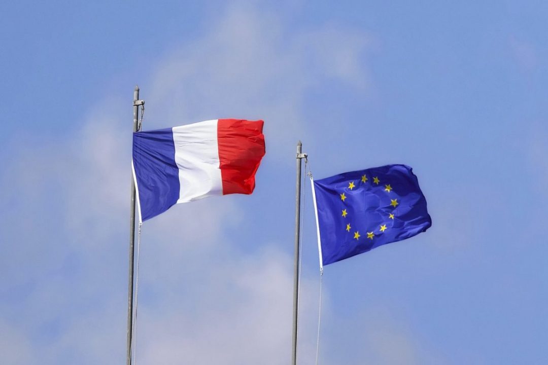Deux drapeaux flottent côte à côte sur un fond de ciel bleu : le drapeau français, et le drapeau européen.