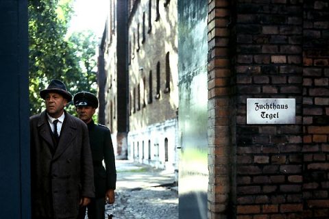 Image tirée du film Berlin Alexanderplatz de Rainer Werner Fassbinder : un homme en chapeau et manteau long sort de la cour d'une prison avec, derrière lui, un gardien et un bâtiment austère