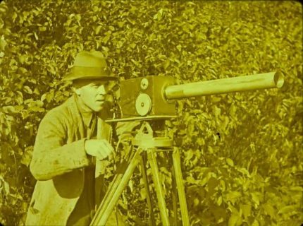 Pellicule teintée en jaune. Un homme manie une caméra avec téléobjectif devant des arbres.