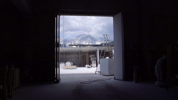 Dans un atelier sombre, une porte est ouvert sur la montagne barrée de carrières de marbre blanc, et lumineuse.