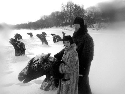 Photo en noir et blanc d'un homme et d'une femme en manteaux de fourrure avec, en arrière plan, des têtes de chevaux enlisés dans la neige