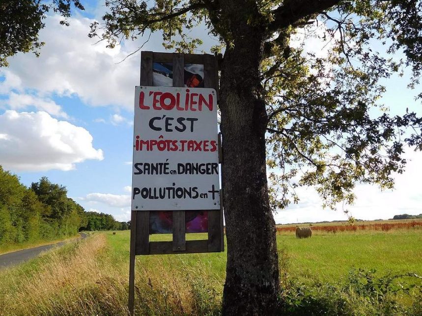 Une pancarte plantée contre un arbre, en bordure d'un champ, sur laquelle est écrit L'éolien c'est impôts, taxes, santé en danger, pollutions en plus