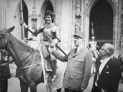Photographie en noir et blanc d'une jeune fille en armure et à cheval, déguisée en Jeanne d'Arc, serrant la main du général de Gaulle, en uniforme, debout à ses côtés