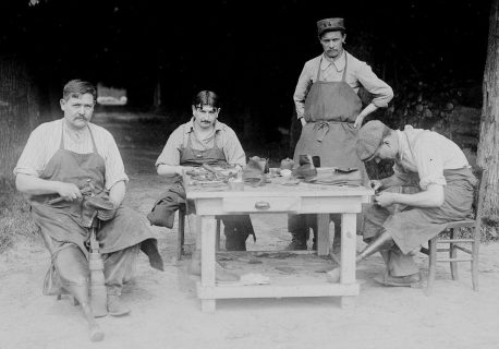Photographie en noir et blanc : trois hommes, amputés d'une jambe, sont assis autour d'une table en plein air et manipulent du cuir ou des chaussures. Un quatrième est debout à côté.