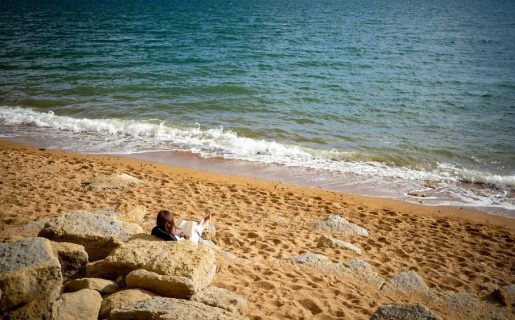 Une femme en train de lire, assise le dos contre des rochers, sur une plage, face à la mer