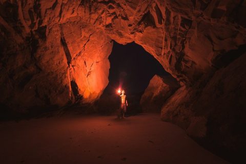 Un homme debout dans une grotte, éclairée par la lueur rouge d'une torche qu'il tient à la main
