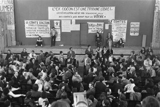 Photographie en noir et blanc : une foule assise dans un théâtre, face à la scène, sur laquelle sont accrochées des banderoles