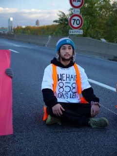 un militant de Dernière rénovation opérant un sit-in sur l'autoroute. Il porte un t-shirt marqué : We have 858 days left, et un gilet orange