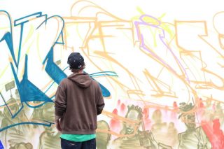 Une personne avec une casquette et un hoodie, de dos, regarde un graffiti sur un mur.