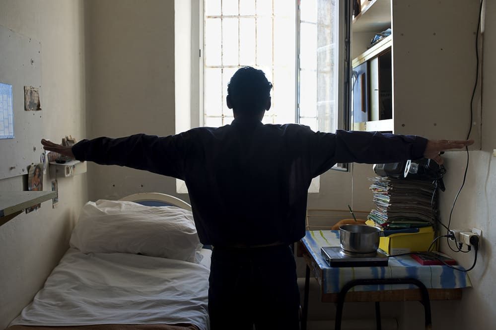Un homme à contre-jour étend les bras sur le côté, touchant presque les murs de sa cellule de prison.