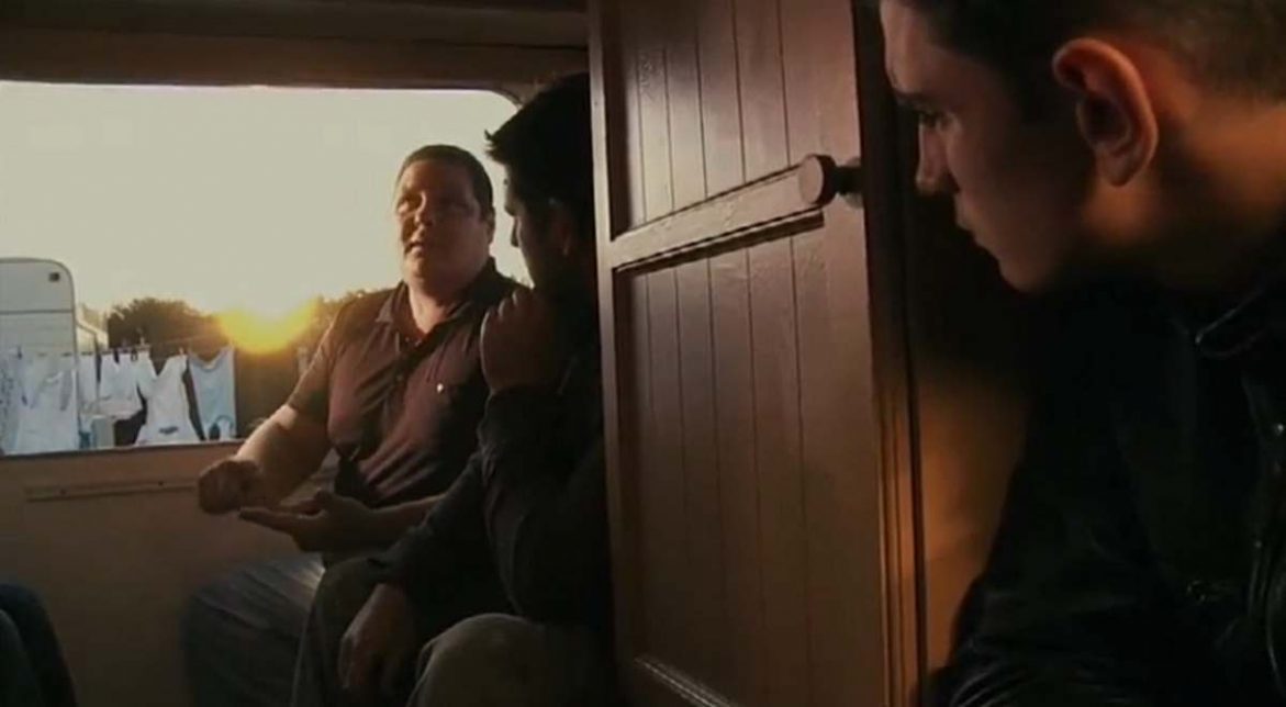 Fred dans sa caravane parle avec un autre homme; Un troisième se tient à la porte. Au second plan le camp, une caravane au soleil couchant et du linge qui sèche sur un fil.