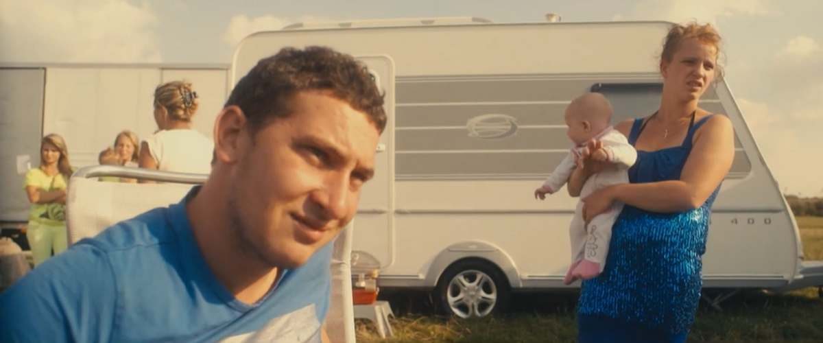 Scène de vie du camp de gitans. Au premier plan, un homme et une femme tenant un bébé dans ses bras. A l'arrière plan une caravane et des femmes qui discutent.