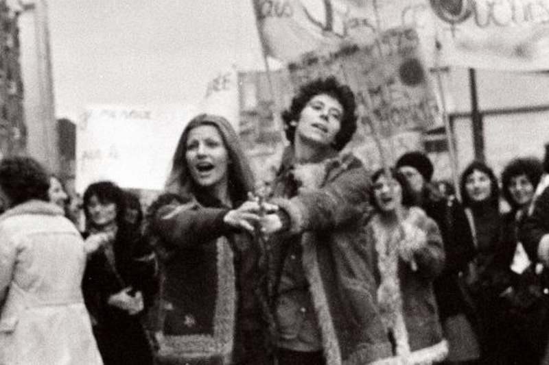 Photo d'archive en noir et blanc montrant une manifestation dans les années 1970. Au premier plan deux personnes dansent, au second plan des manifestantes avec des banderoles.