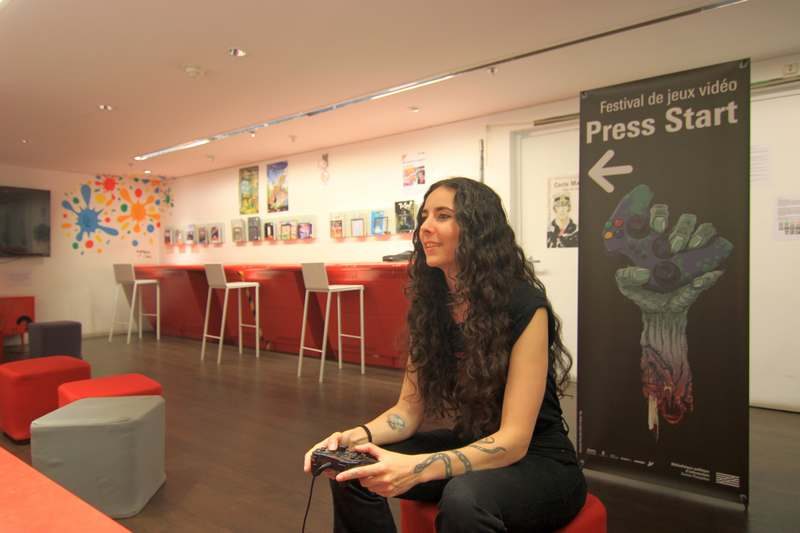 Taous Merakchi, marraine de l'édition 2024 du festival de jeu vidéo Press Start, au salon de jeux vidéo de la Bpi. Elle tient une manette de jeu.