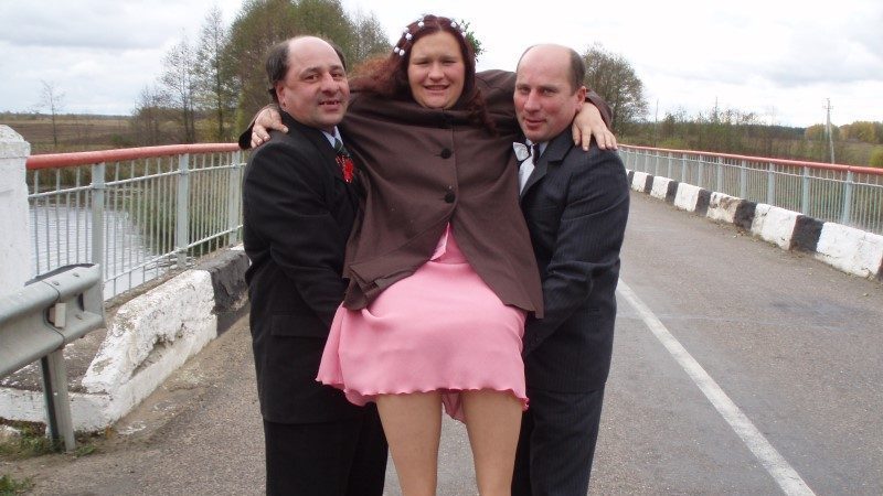 Une femme en robe rose est portée par deux hommes en costume, au cours d'un mariage. Elle a ses bras autour de leurs nuques.