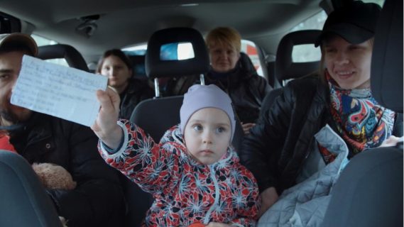 Des gens sont dans une voiture, entassés. Parmi les passagers et passagères, une fillette tend un papier devant elle.
