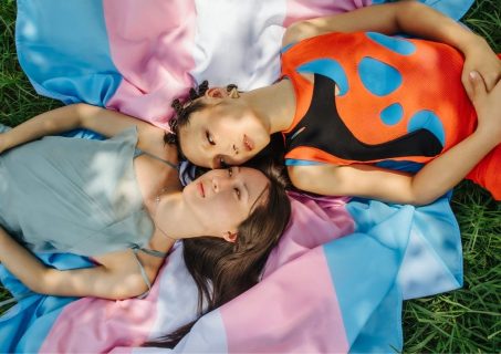 Photo couleur : vue en plongée sur 2 femmes allongées dans l'herbe, sur un drapeau transgenre bleu ciel, rose et blanc.