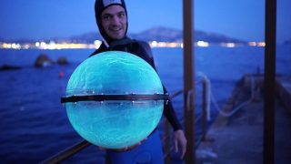 Jeremy Bruguidou en tenue de plongée, une de ses sphères bioluminescentes à la main. La sphère est de la taille de son torse