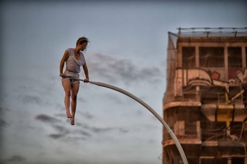 L'artiste Chloé Moglia, pieds nus, en tenue blanche, se tient en suspension sur un barre d'appui blanche courbée. En arrière plan, un bâtiment en friche apparaît