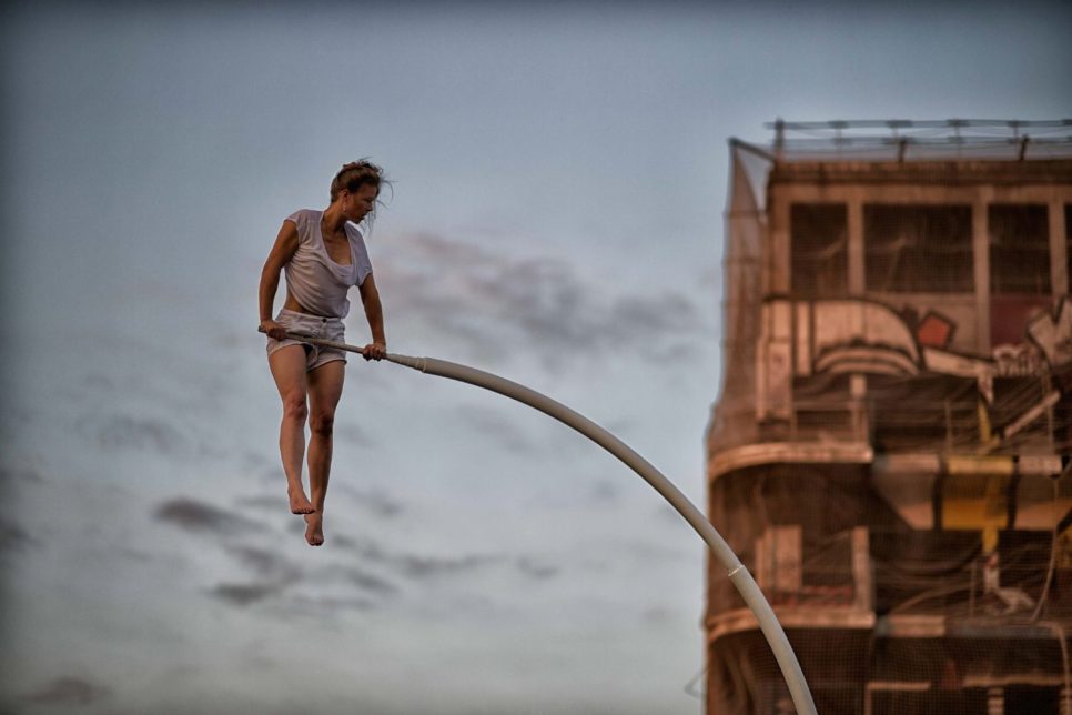 L'artiste Chloé Moglia, pieds nus, en tenue blanche, se tient en suspension sur un barre d'appui blanche courbée. En arrière plan, un bâtiment en friche apparaît