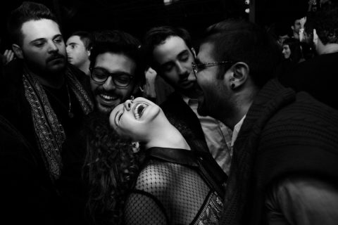 Photographie en noir et blanc de la photographe libanaise Myriam Boulos, issue de sa série Nighshift réalisée en 2015. Scène de fête, dans un club de Beyrouth, le B018. Gros plan sur une jeune femme qui rit, gorge déployée, inclinée en arrière. Elle est entourée d’hommes qui rient et dansent.